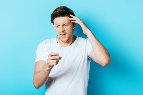 Angry man looking at smartphone on blue background - Photo, Image