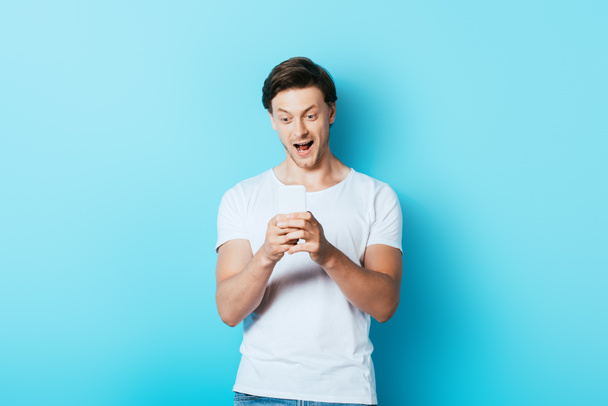 Excited man in white t-shirt using smartphone on blue background - Photo, image