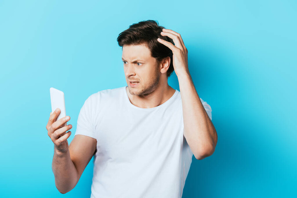 Στοχαστικός άνθρωπος με το χέρι κοντά στο κεφάλι χρησιμοποιώντας smartphone σε μπλε φόντο - Φωτογραφία, εικόνα