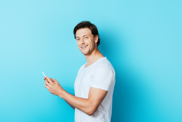 Young man looking at camera while holding smartphone on blue background - Foto, Bild