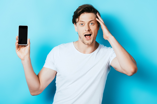Excited man showing smartphone with blank screen and looking at camera on blue background - Fotó, kép