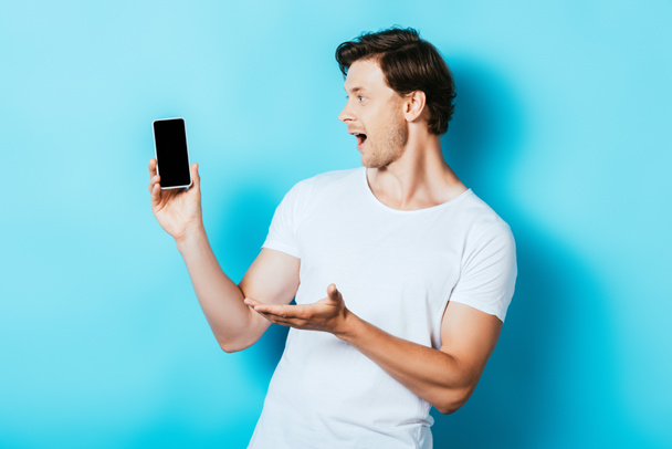 Surprised man pointing with hand while holding smartphone with blank screen on blue background - Фото, изображение