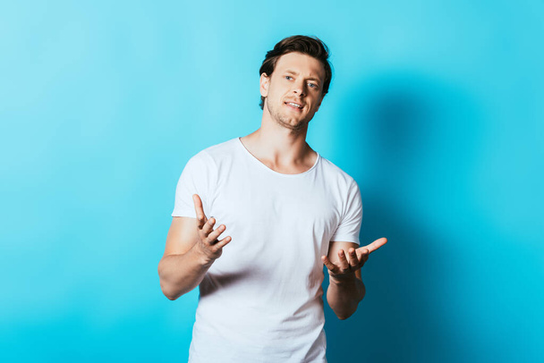Homme confiant en t-shirt blanc pointant les mains vers la caméra sur fond bleu - Photo, image