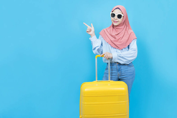 Portrait de jeune belle femme musulmane asiatique en robe traditionnelle avec valise jaune ou bagage isolé sur fond bleu, Vacances d'été, vacances et concept de voyage - Photo, image