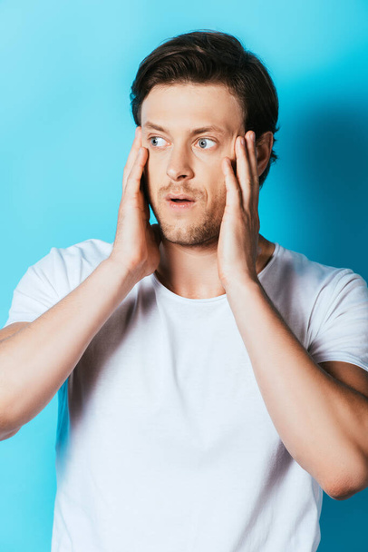 Worried man in white t-shirt with hands near face on blue background - Foto, Bild