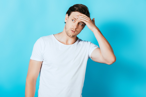 Fiatal férfi fehér pólóban megható homlok, miközben félrenéz a kék háttér - Fotó, kép