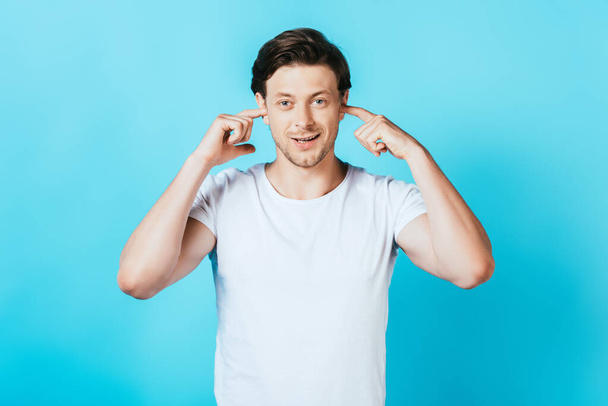 Young man looking at camera while covering ears with fingers on blue background - Foto, Imagem