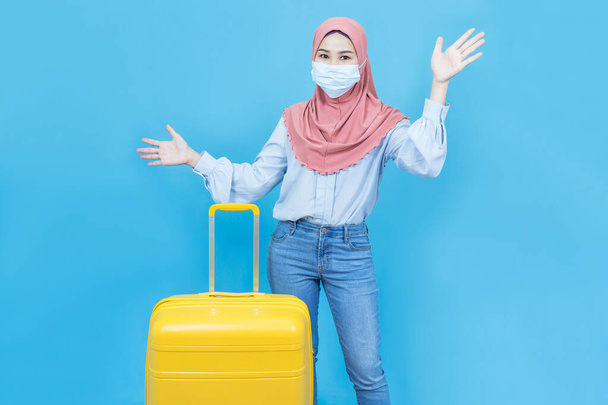 Portrait of young beautiful asian muslim woman wearing medical face mask in traditional dress with yellow suitcase or luggage  isolated on blue background,Summer holidays, vacation and travel concept - Photo, image