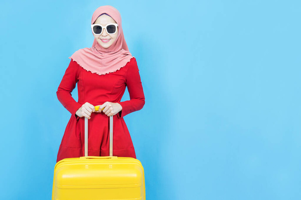 Retrato de joven hermosa mujer musulmana asiática en vestido tradicional rojo con gafas de sol con maleta amarilla o equipaje aislado sobre fondo azul, vacaciones de verano, vacaciones y concepto de viaje - Foto, Imagen