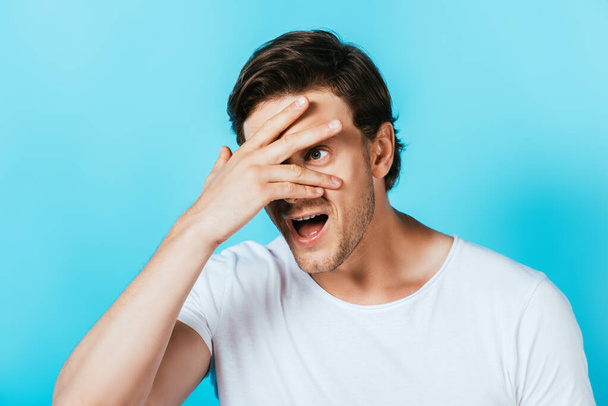 Young man looking away while covering face with hand isolated on blue  - Foto, Imagen