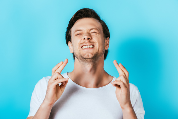 Young man in white t-shirt with crossed fingers on blue background - Photo, Image