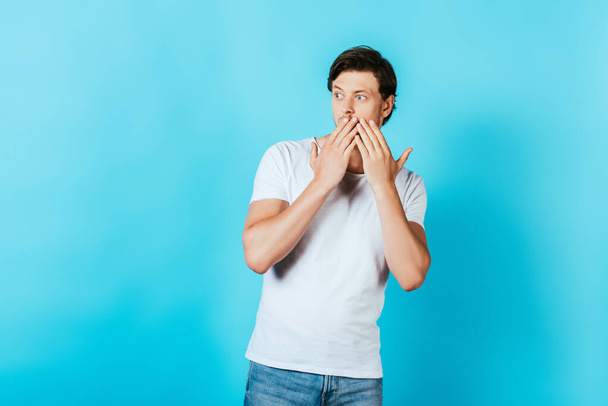 Έκπληκτος άνθρωπος σε λευκό t-shirt που καλύπτει το στόμα με τα χέρια σε μπλε φόντο - Φωτογραφία, εικόνα