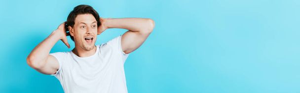 Panoramic shot of ecited man with hands near head looking away on blue background - Фото, изображение