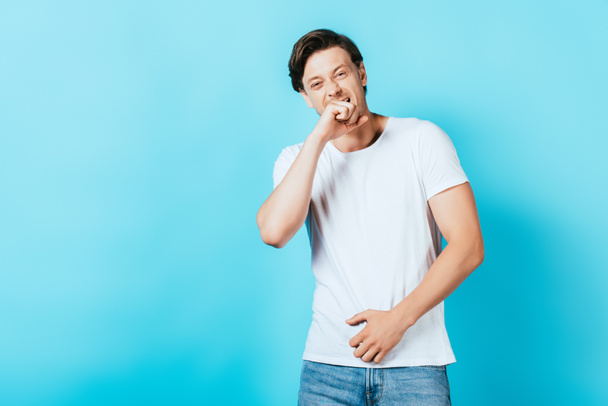 Γέλιο άνθρωπος με το χέρι κοντά στο στόμα κοιτάζοντας κάμερα σε μπλε φόντο - Φωτογραφία, εικόνα