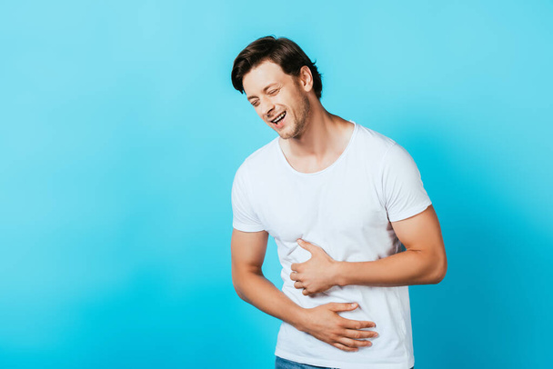 Young man in white t-shirt laughing and touching belly on blue background - Photo, image