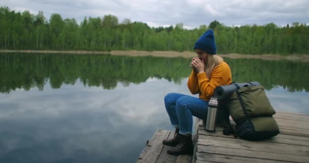 Nainen, jolla on hattu ja reppu, istuu metsäjärven puulaiturilla vuorilla ja juo kuumia juomia teetä tai kahvia termospullosta.. - Materiaali, video