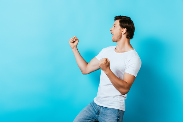 Hombre excitado en camiseta blanca mostrando sí gesto sobre fondo azul - Foto, Imagen
