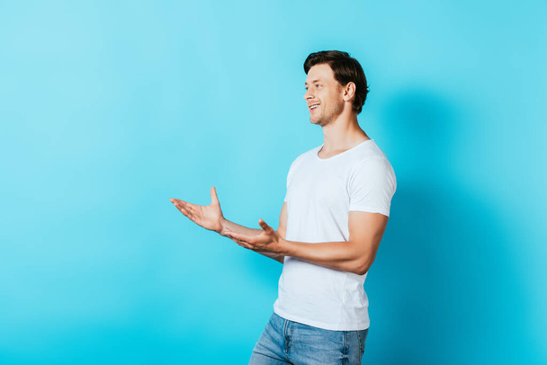 Fiatal férfi fehér pólóban, mutatva a kezével, és félrenéz a kék háttér - Fotó, kép