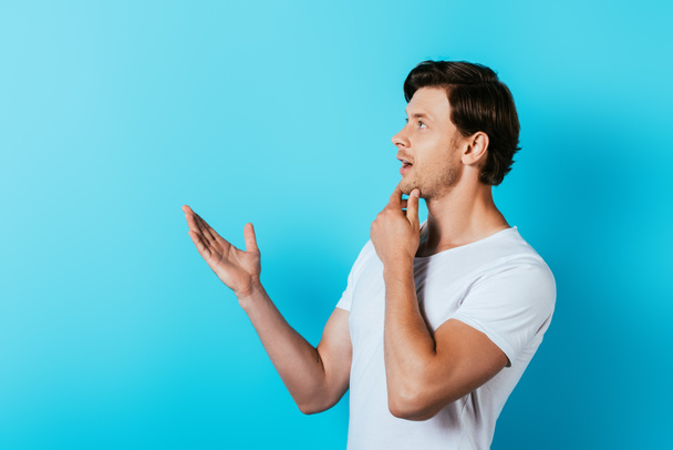 Pensive man in white t-shirt pointing with hand on blue background - Fotografie, Obrázek