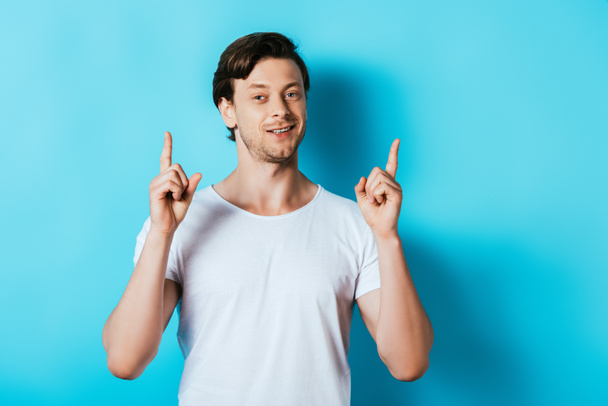 Young man in white t-shirt pointing with fingers up on blue background - Foto, afbeelding
