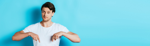 Panoramic shot of young man in white t-shirt pointing with fingers on blue background - Zdjęcie, obraz
