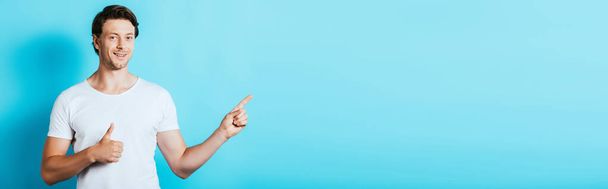 Horizontal concept of young man showing thumb up and pointing with finger on blue background - Photo, Image