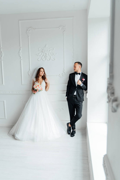 De eerste ontmoeting van de bruidegom in een zwart pak en de bruid in een witte trouwjurk met een boeket in het interieur van een fotostudio op een witte en zwarte achtergrond - Foto, afbeelding