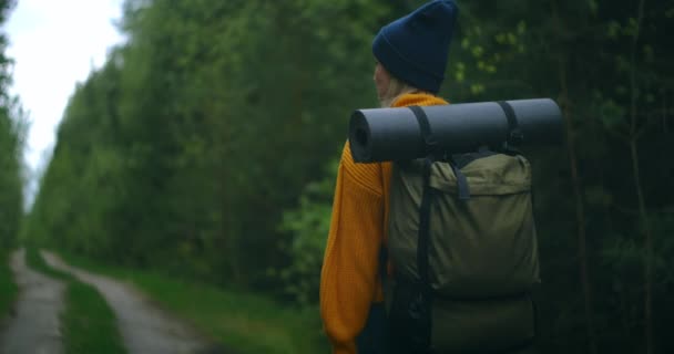 背中の下と側からの眺め:女性は黄色のセーターとバックパック付きの森林道路上の帽子で徒歩で旅行し、木や自然を見ています。一人旅で。自由の概念  - 映像、動画