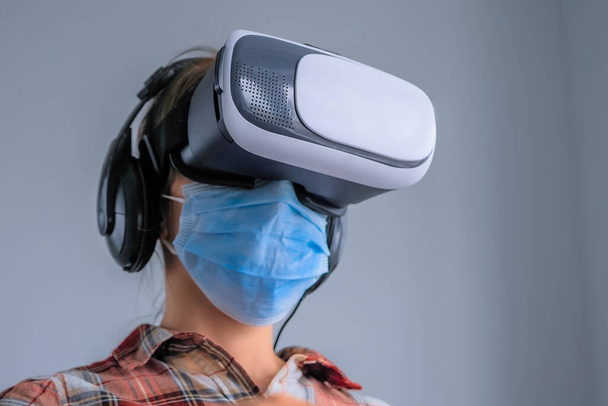 Giovane donna con maschera medica, auricolare realtà virtuale contro muro grigio - Foto, immagini