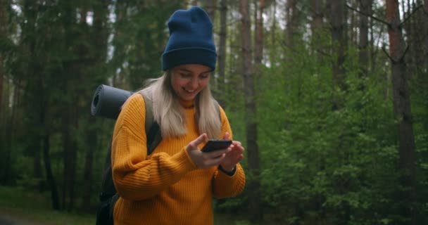 Kobieta w żółtym swetrze i kapeluszu z plecakiem trzyma telefon komórkowy i wpisuje wiadomość. Tapping palce na ekranie i uśmiech. Podróżnik w lesie na drodze używa smartfona - Materiał filmowy, wideo