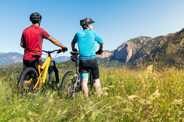 vista posterior de dos hombres observando el paisaje y las montañas de pie junto a su bicicleta de montaña en el campo, concepto de deporte y estilo de vida saludable en la naturaleza, espacio de copia para el texto - Foto, Imagen