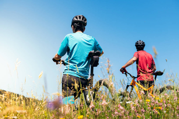 back view of two cyclists standing next to their mountain bikes in a field with flowers, concept of sport with friends and healthy lifestyle in nature, copy space for text - Photo, image