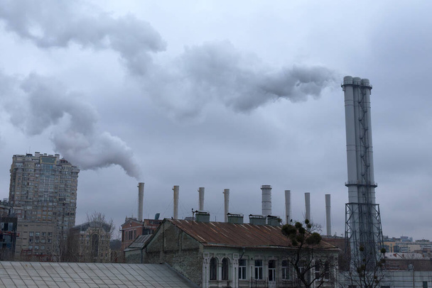 Dym pochodzi z komina przemysłowego nad dachami. Niebo jest pokryte szarymi chmurami. Zanieczyszczenie środowiska. - Zdjęcie, obraz
