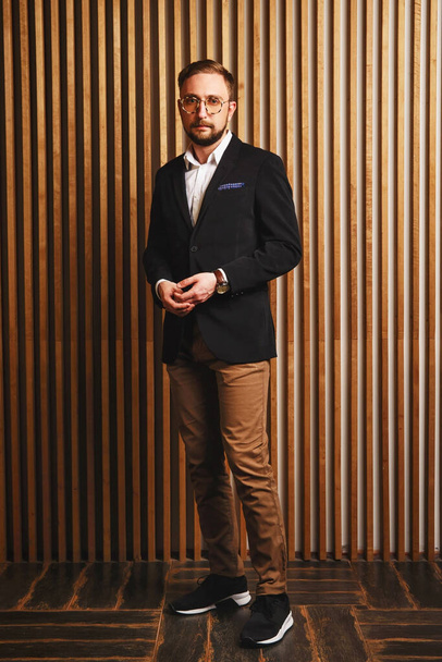 Portrait of young beautiful fashionable man in glasses, brown blaizer and trousers against wooden wall . Hipster style guy looking at camera serious - Foto, afbeelding