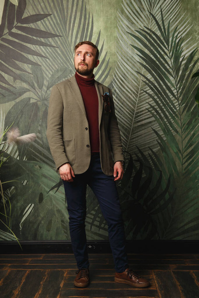 Portrait of young beautiful fashionable man, in olive jacket blaizer and blue trousers against wallpaper with greenery. Hipster style guy looking aside and thinking - Φωτογραφία, εικόνα