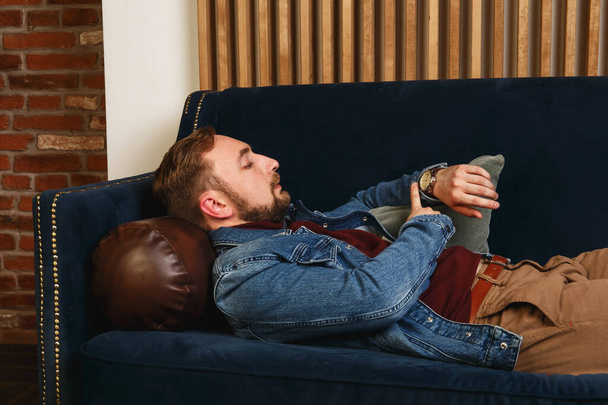 Beautiful elegant fashionable young man in a blue jacket lying on a sofa in modern appartment and looks at his watch - Foto, Imagen