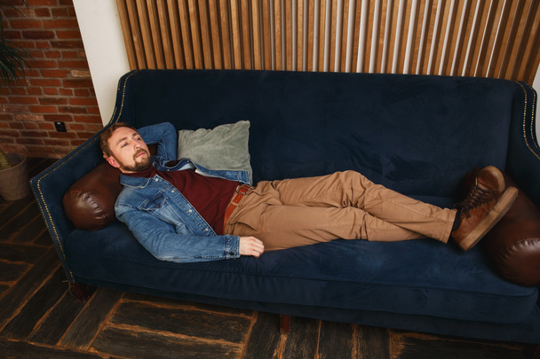 Desde arriba vista de un hermoso hombre joven elegante de moda en una chaqueta azul acostado en un sofá en el compartimiento moderno y pensando - Foto, Imagen