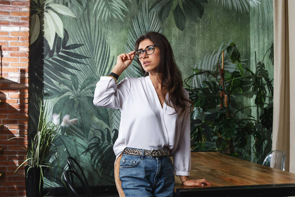 Sexy young girl in glasses white shirt and blue jeans standing in modern appartment by wooden table, looks away thoughtfully - Foto, afbeelding
