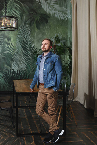 Portrait of handsome bearded man in stylish casual clothes blue denim jacket and beige pants standing in modern appartment interior by wooden table looking aside and thinking - Valokuva, kuva