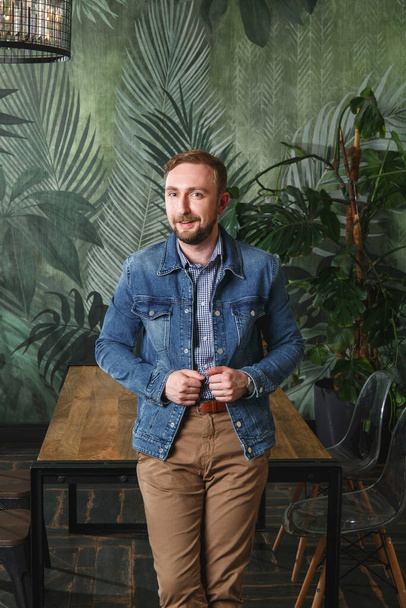 Portrait of handsome man in casual clothes blue denim jacket and beige pants standing in modern appartment by wooden table looking at camera and smiling - Foto, afbeelding