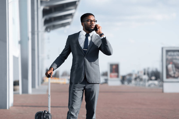 Entrepreneur africain parlant sur téléphone portable debout avec valise à l'extérieur - Photo, image