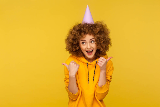 Portrait of joyful curly-haired hipster woman with funny cone hat pointing advertising area and shouting in amazement, showing copy space for commercial text about holidays, sales, crazy event - Fotoğraf, Görsel