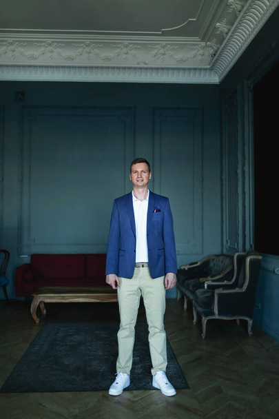 Handsome stylish tall man in blue jacket and beige trousers standing in a dark room with classic interior and looking at camera and smiling - Photo, Image
