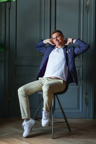 Handsome stylish man in blue jacket and white shirt sitting on chair in a room with classic interior, looking at camera and smiling - Фото, зображення