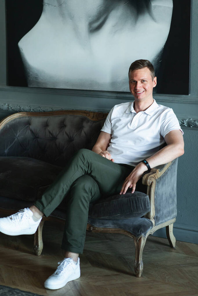 Handsome stylish tall man in in a polo, green pants and white sneakers sitting on soft sofa in a room with classic interior and looking at camera and smiling - Foto, immagini