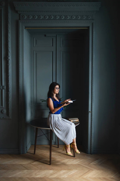Stylish beautiful longhaired woman a blue shirt and skirt sitting in glasses on round wooden table in appartment interior and reading book. - Foto, Imagen