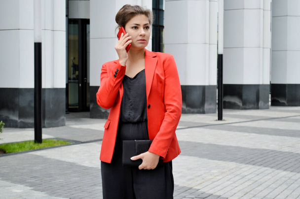 Красивая деловая женщина на фоне современного офиса. Девушка в черном комбинезоне и красной куртке разговаривает по телефону возле бизнес-центра - Фото, изображение