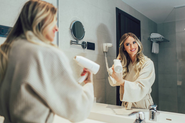 Beautiful women enjoying in hotel room.Young happy smiling woman wearing bathrobe using hair dryer in bathroom. - Fotó, kép