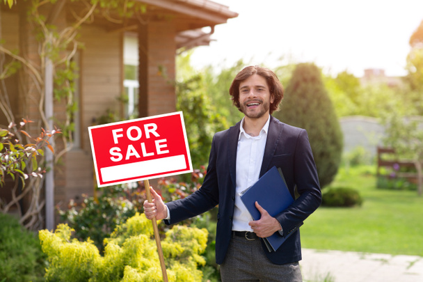 Happy real estate agent holding FOR SALE sign near residential building outdoors - Photo, Image