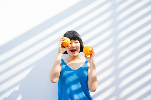 alegre feliz hermosa bob corte de pelo asiático mujer en azul camiseta de tanque de celebrar dos naranjas tropical fruta en las manos sobre fondo blanco. - Foto, Imagen